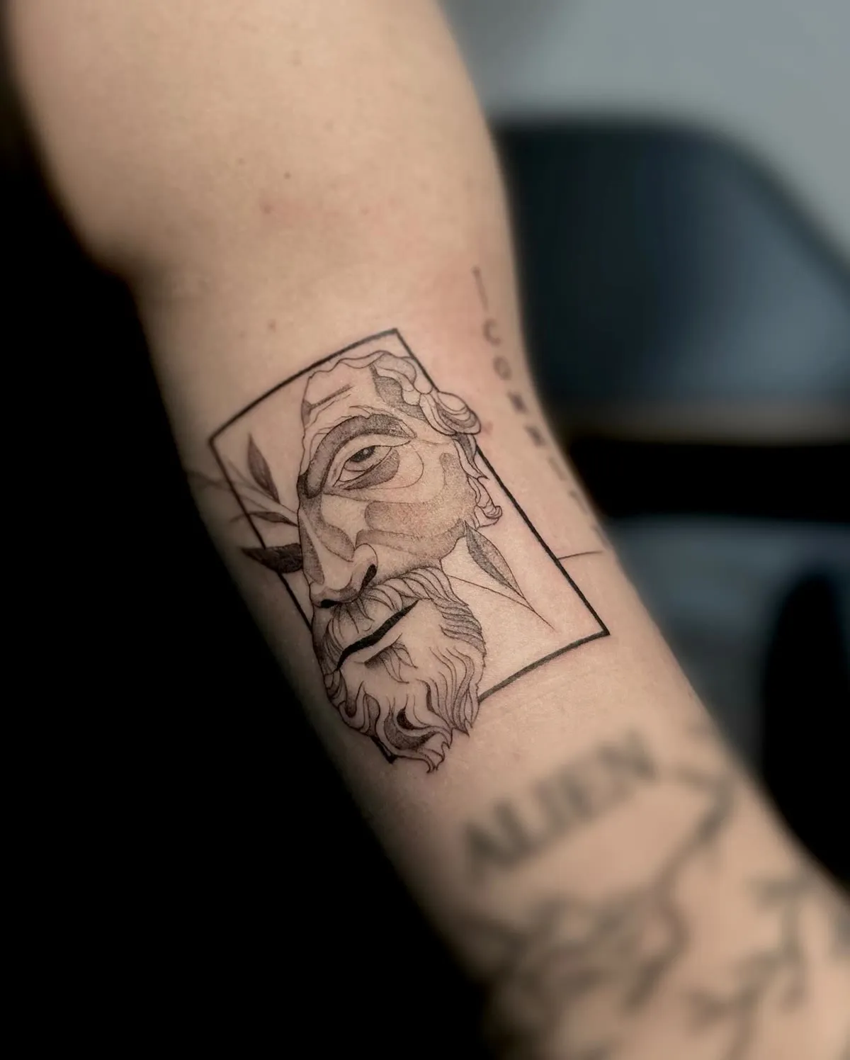 A bearded face inside a drawn frame, etched on a forearm above a line of lettering.